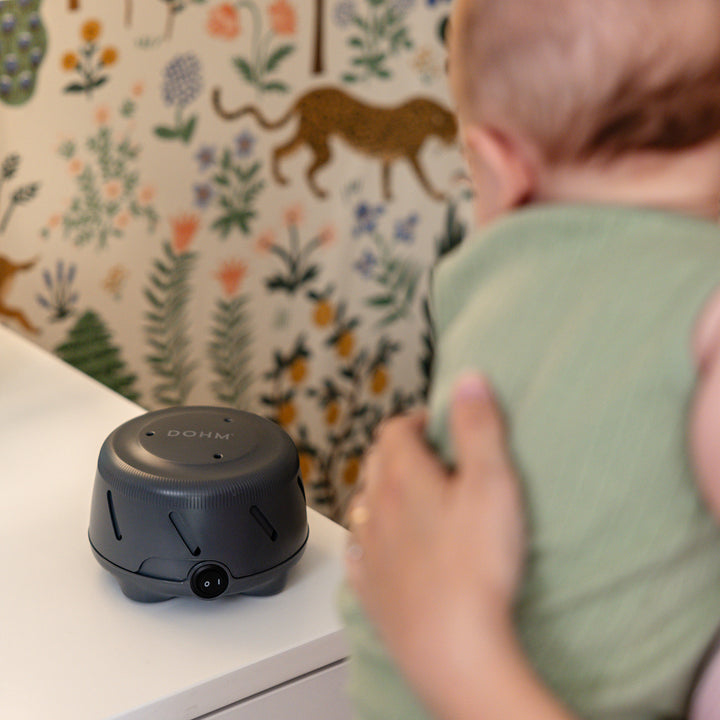 Charcoal Dohm Uno white noise machine on a dresser as a parent holds a baby in a floral nursery