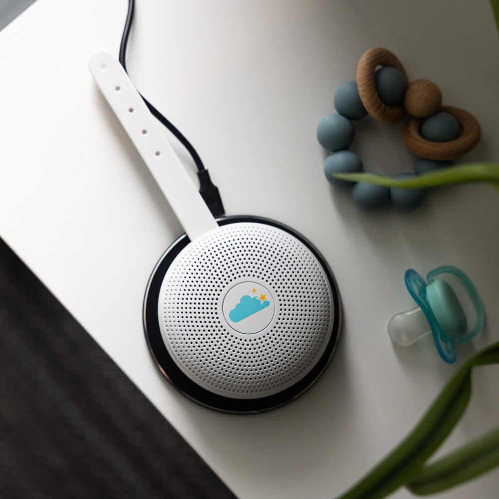 Top-down view of Hushh travel sound machine with white strap and cloud logo on a white surface beside a pacifier and teething toy