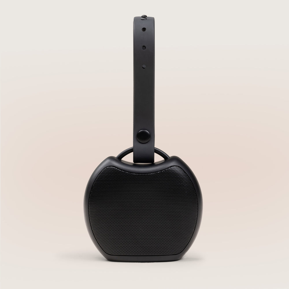 Front view of Rohm Plus portable sound machine showing black speaker grille and adjustable strap on a beige background