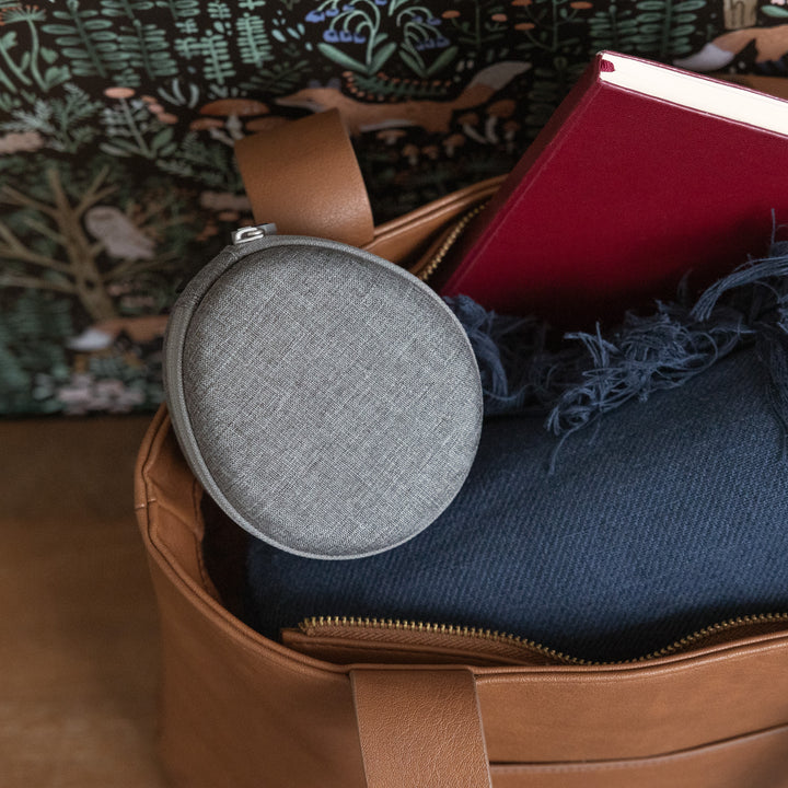Top view of Travel Case showing grey linen fabric, round zippered shape, and small carrying strap tucked inside a brown tote bag