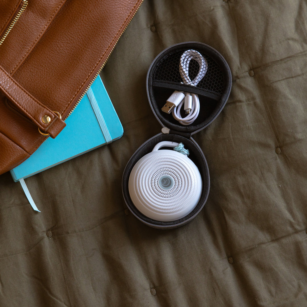 Top view of travel case with Hushh white sound machine and USB cable inside on a brown quilt beside a leather bag and teal notebook