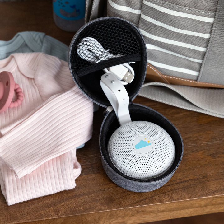 Open travel case with white Hushh sound machine and USB cable in mesh pocket on wooden table beside baby clothes