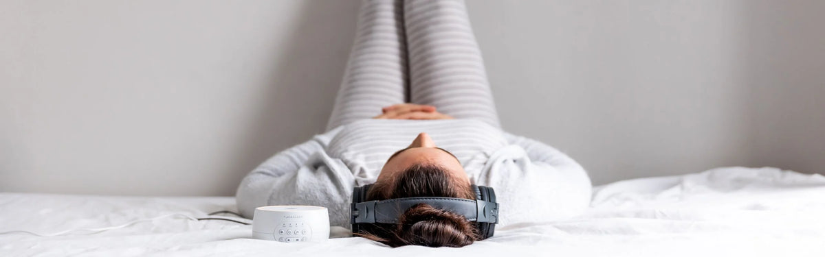 Woman lying on a bed wearing headphones with legs rested against the wall beside a white Yogasleep sound machine