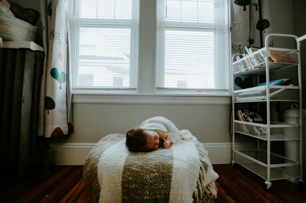 Infant sleeping peacefully on a cushioned ottoman blanket beneath a sunny nursery window