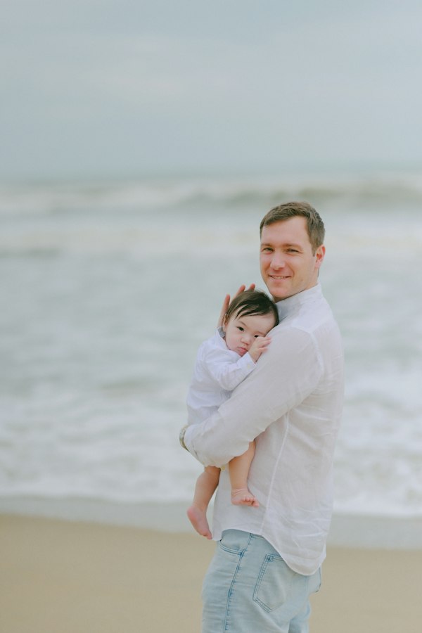 Father in a white shirt holding his baby in his arms standing on a sandy ocean beach