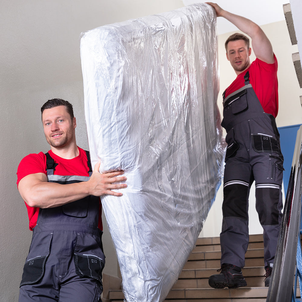 Two movers carrying a plastic-wrapped mattress down an indoor stairwell during delivery